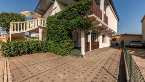 Una casa de dos pisos con entrada pavimentada, una escalera con barandilla y un balcón con flores rojas.