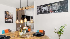 A dining area with a wooden table set for four, surrounded by chairs with orange cushions. There are hanging light fixtures above the table, and a framed picture on the wall to the right. - MOLA Apartments-Style & Flair (Wetter (Ruhr))