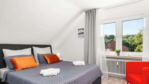 A modern bedroom with a large bed, a red chair, and a window with a view of greenery and buildings. - MOLA Apartments-Style & Flair (Wetter (Ruhr))