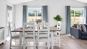 A bright, spacious room with a dining table and chairs, large windows with curtains, a grey sofa, and a potted plant. - Poppy, 5 Roadford Lakes Lodges  -  a lodge that sleeps 4 guests  in 2 bedrooms (Roadford Lake)