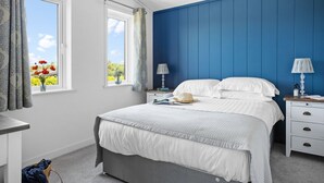 A bedroom with a bed, bedside tables, a nightstand, a window with curtains, and a view of greenery outside. - Poppy, 5 Roadford Lakes Lodges  -  a lodge that sleeps 4 guests  in 2 bedrooms (Roadford Lake)