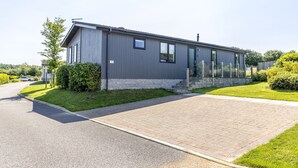 A modern single-story building with a grey exterior, a stone base, and a paved driveway leading to a gated entrance. - Poppy, 5 Roadford Lakes Lodges  -  a lodge that sleeps 4 guests  in 2 bedrooms (Roadford Lake)