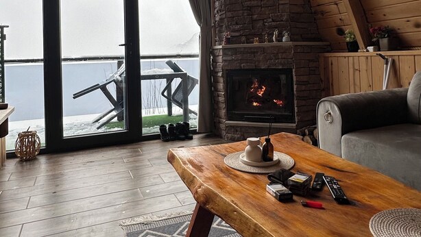 Ein gemütliches Blockhaus-Interieur mit Kamin aus Stein, Holzbalkendecke und großem Fenster mit Blick auf eine verschneite Landschaft.