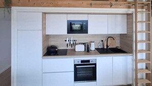 Cocina compacta con horno empotrado, microondas y lavabo, vigas de madera y zona superior tipo altillo.