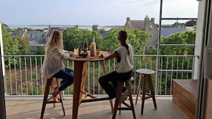 Dos personas sentadas en una mesa de madera en un balcón con vistas a un cuerpo de agua y edificios a lo lejos.