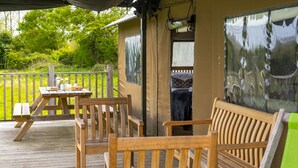 Un terrazzo in legno con tavolo e sedie, una zona riparata con una panchina e vista sul verde.