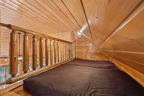 A room with a sloped ceiling, wooden walls, a window with a metal grill, and a mattress on the floor.