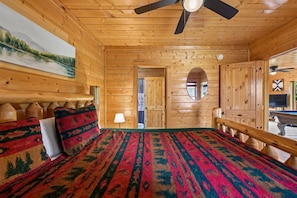 Une pièce de style cabane en bois, avec un lit, un ventilateur de plafond, un tableau et une porte donnant sur une autre pièce.