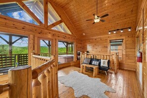 L’intérieur d’un chalet en bois, avec un plafond en bois, un ventilateur de plafond, un canapé avec des coussins, un tapis blanc et une vue sur l’extérieur.