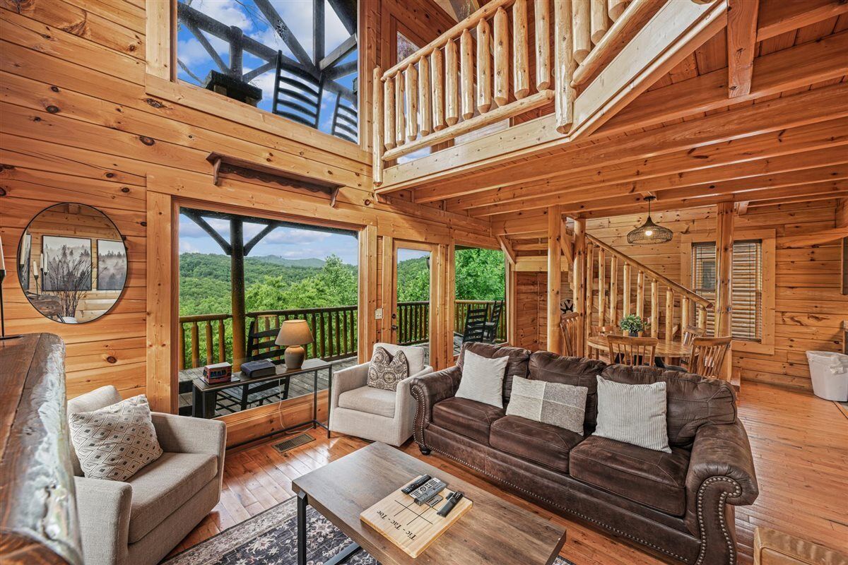 A wooden cabin interior with a living area, a staircase, and large windows overlooking a forested landscape.