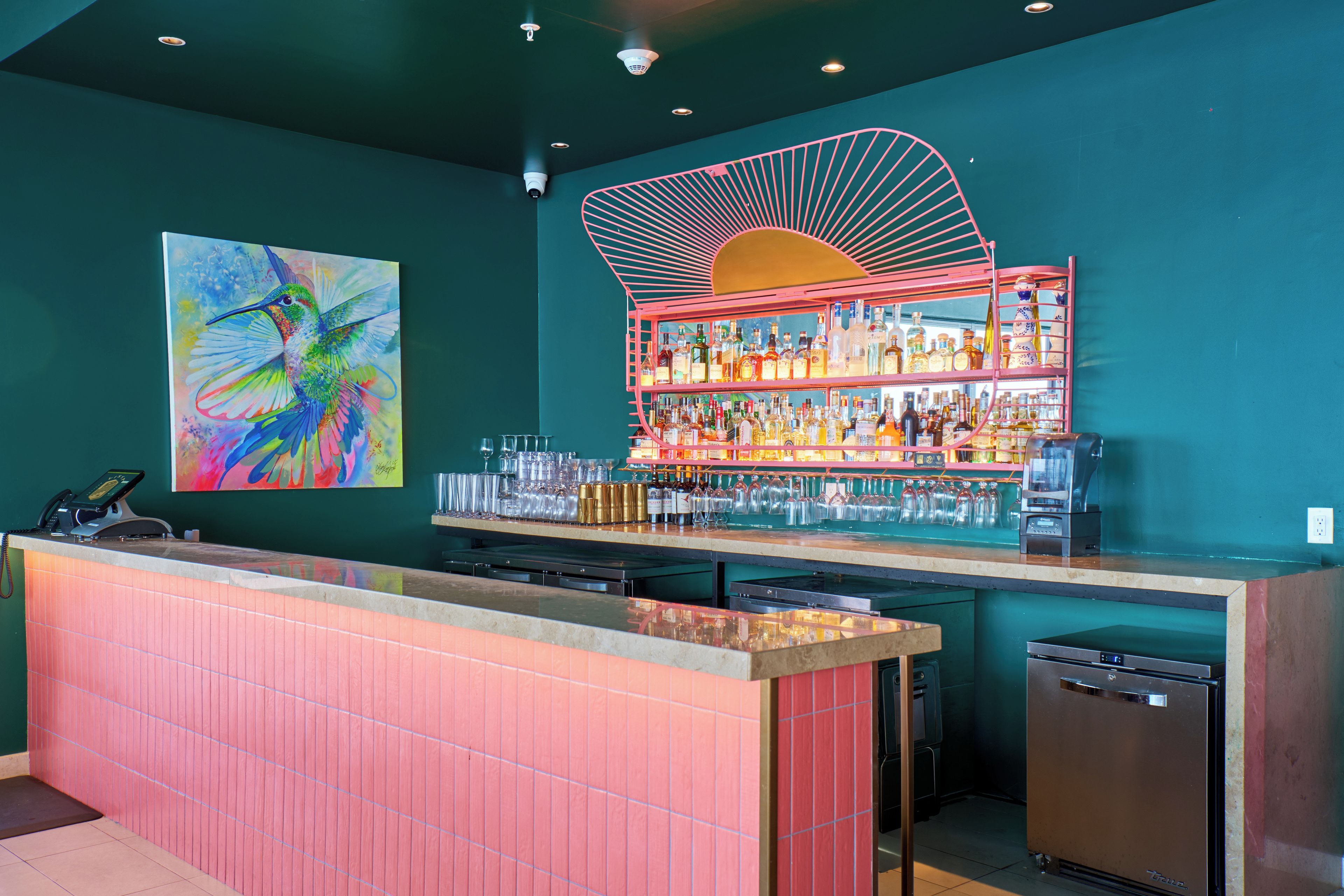 Una zona de bar con mostrador, estantes llenos de botellas, vasos y un cuadro colorido de un pájaro.