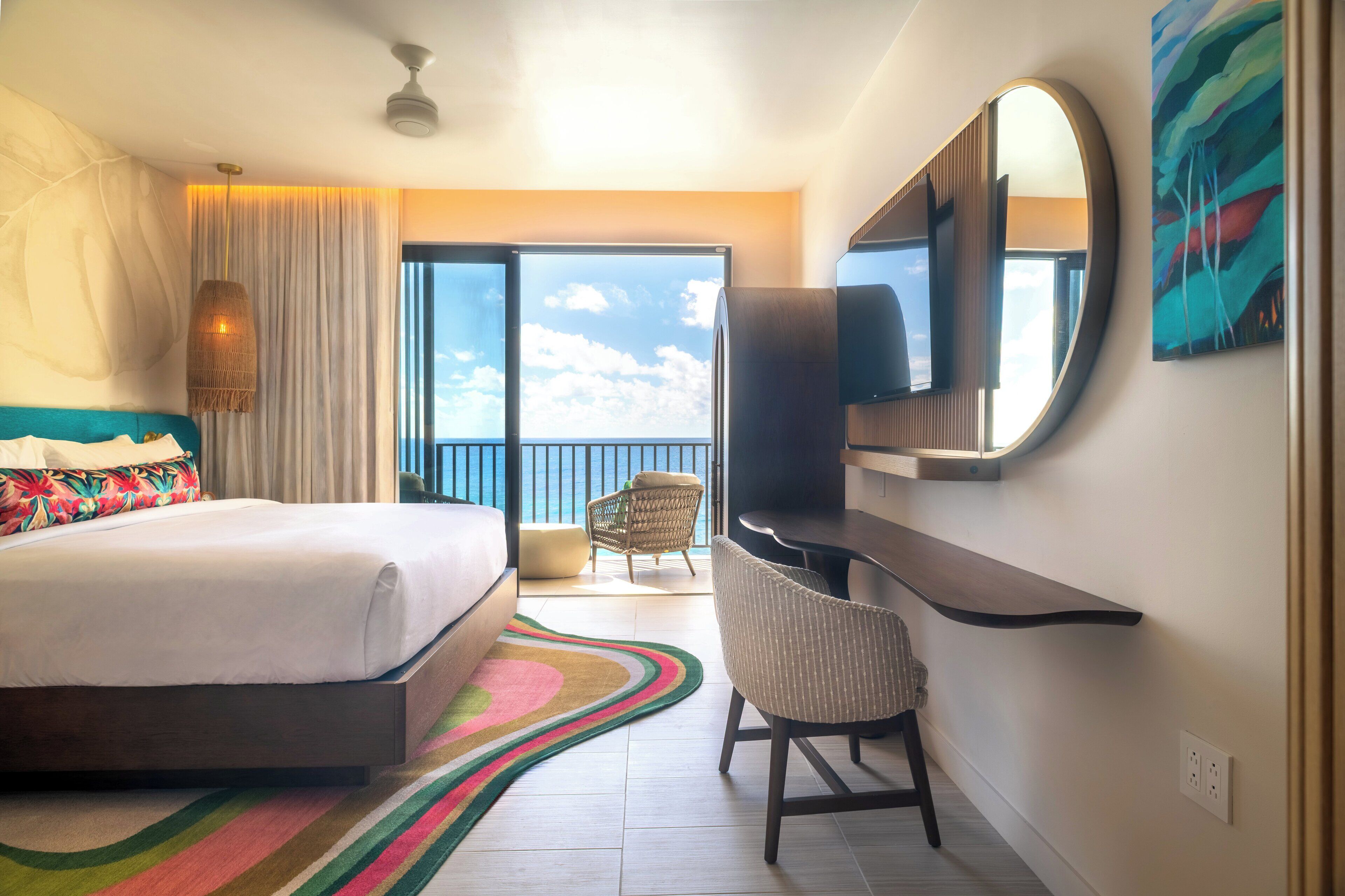 Quarto de hotel moderno com uma cama grande, uma escrivaninha, uma cadeira, uma TV e uma varanda com vista para o céu.