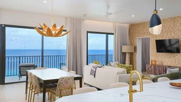 A modern living room with a balcony overlooking the sea, a dining area with a table and chairs, and a kitchen with a marble countertop.