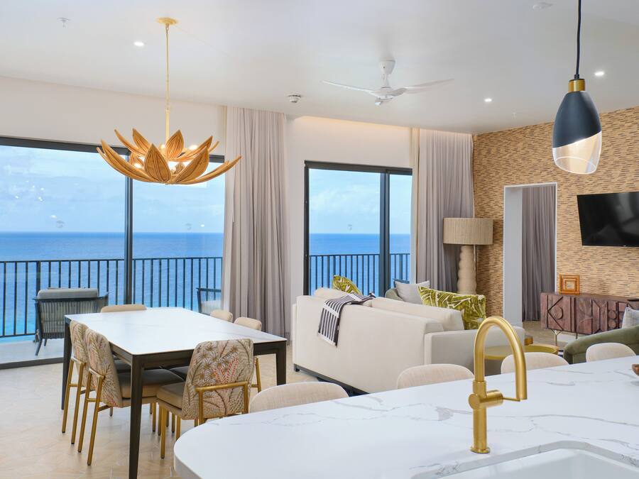 A modern living room with a balcony overlooking the sea, a dining area with a table and chairs, and a kitchen with a marble countertop.
