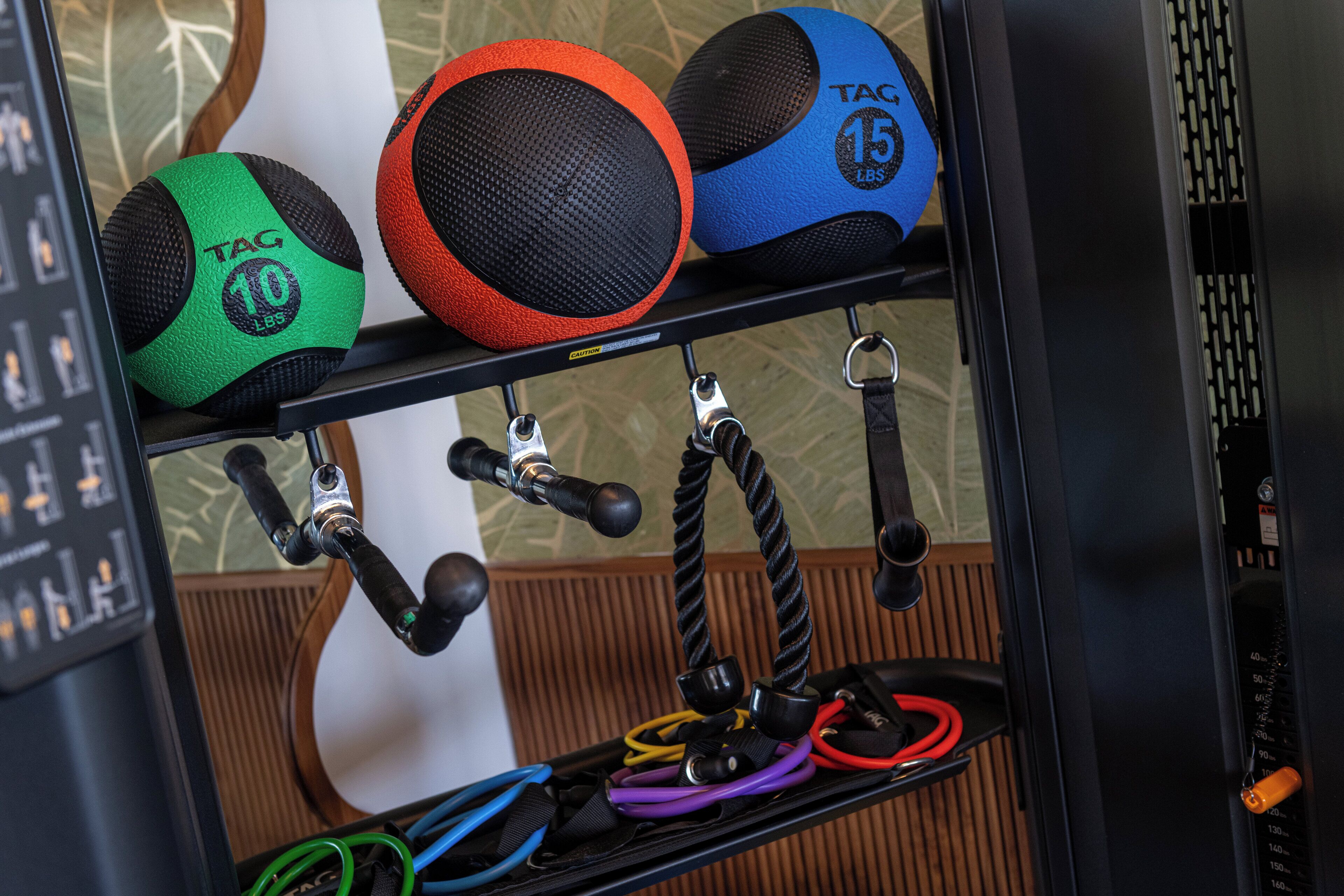 Un estante con equipo de ejercicio, incluyendo una pelota medicinal TAG de 10 libras color verde y negro, una pelota medicinal roja y negra, y una pelota medicinal TAG de 15 libras color azul.