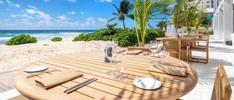 A round wooden table set for dining with glassware and cutlery, overlooking a beach with palm trees and ocean views.