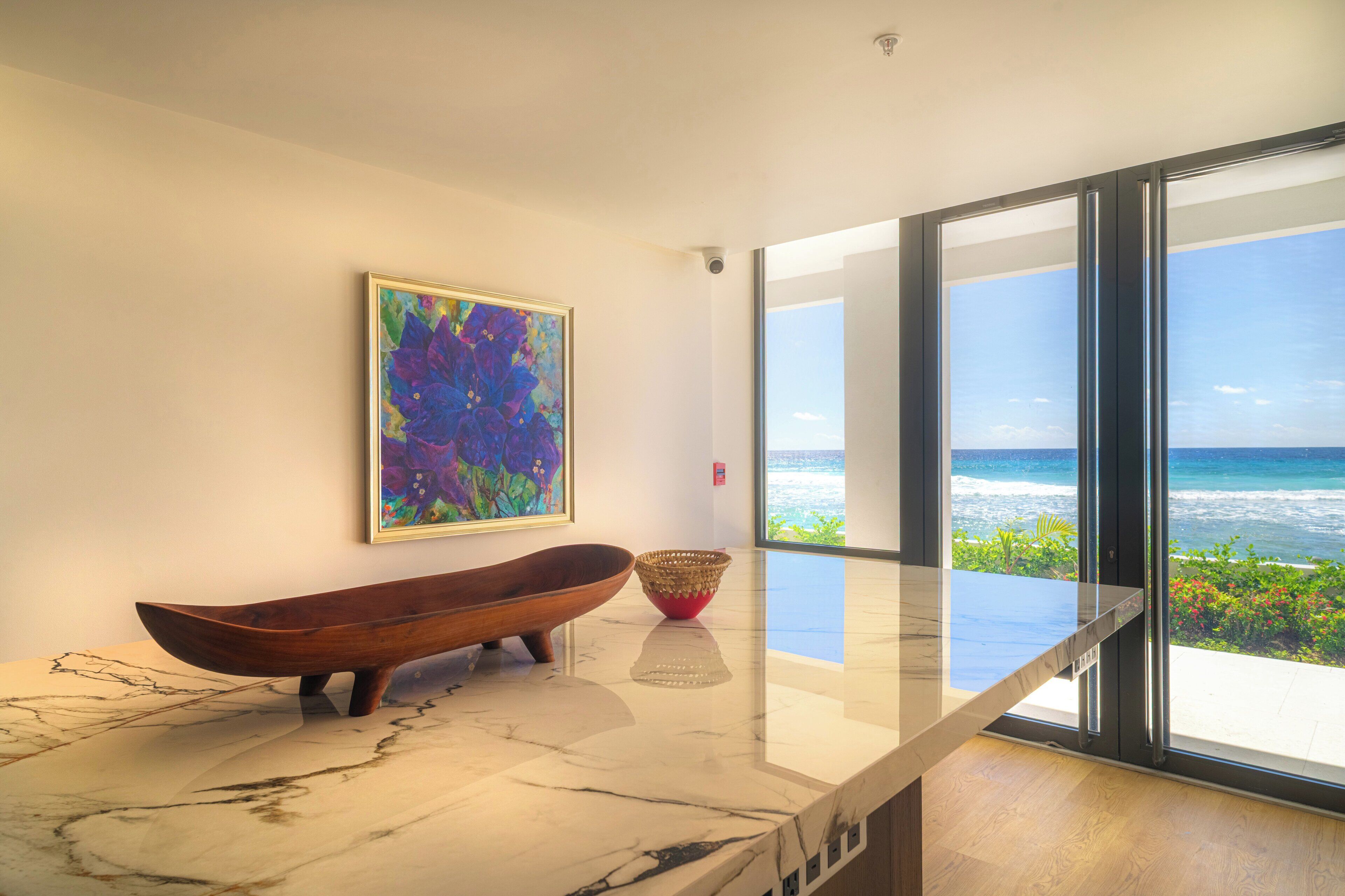 A modern interior with a large window overlooking the ocean, a wooden boat-shaped bowl, and a colorful abstract painting on the wall.