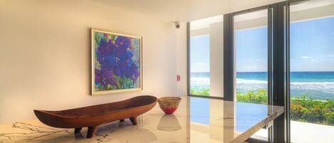 A modern interior with a large window overlooking the ocean, a wooden boat-shaped bowl, and a colorful abstract painting on the wall.