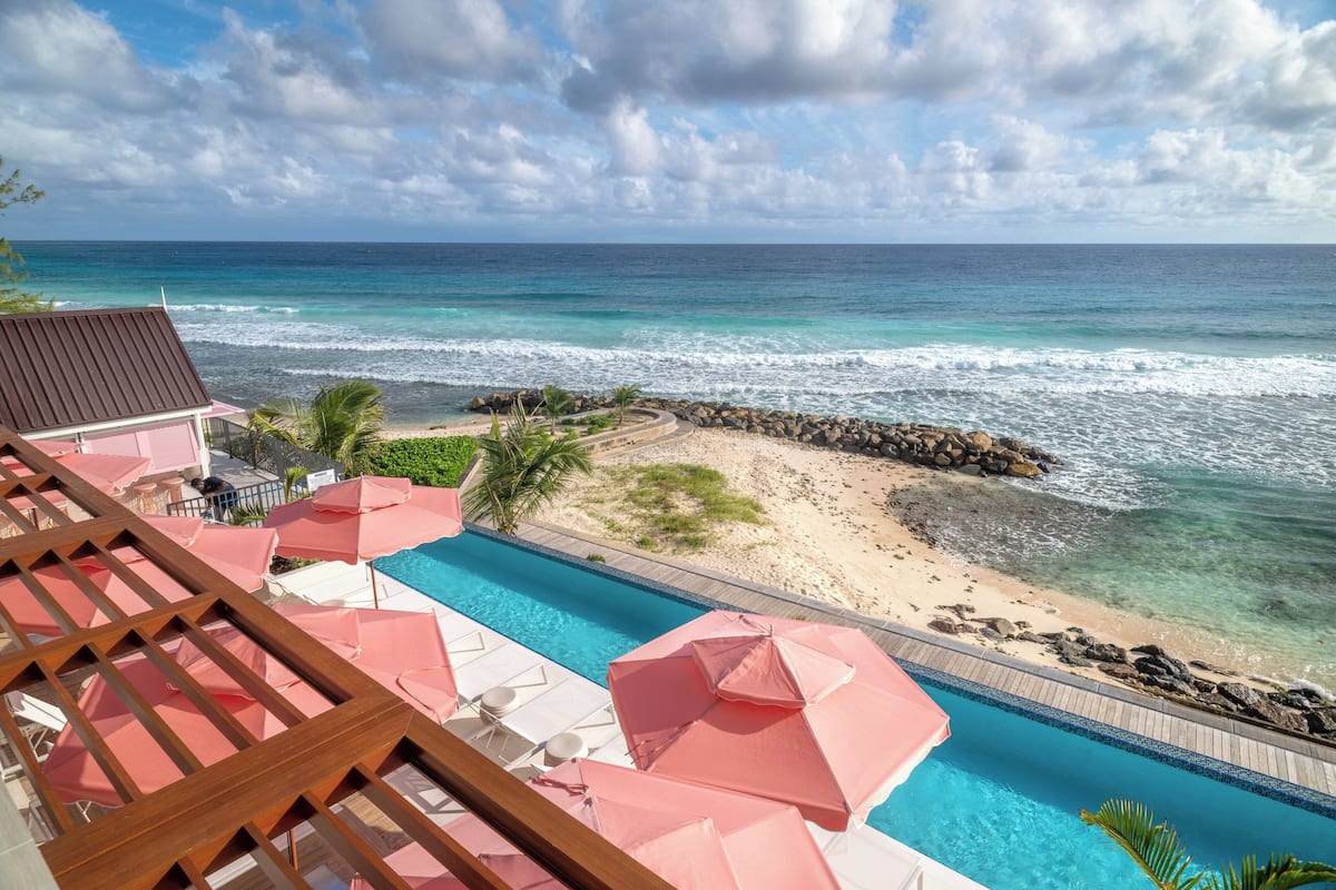 A pool area with pink umbrellas, a view of the ocean, and a sandy beach with rocks.
