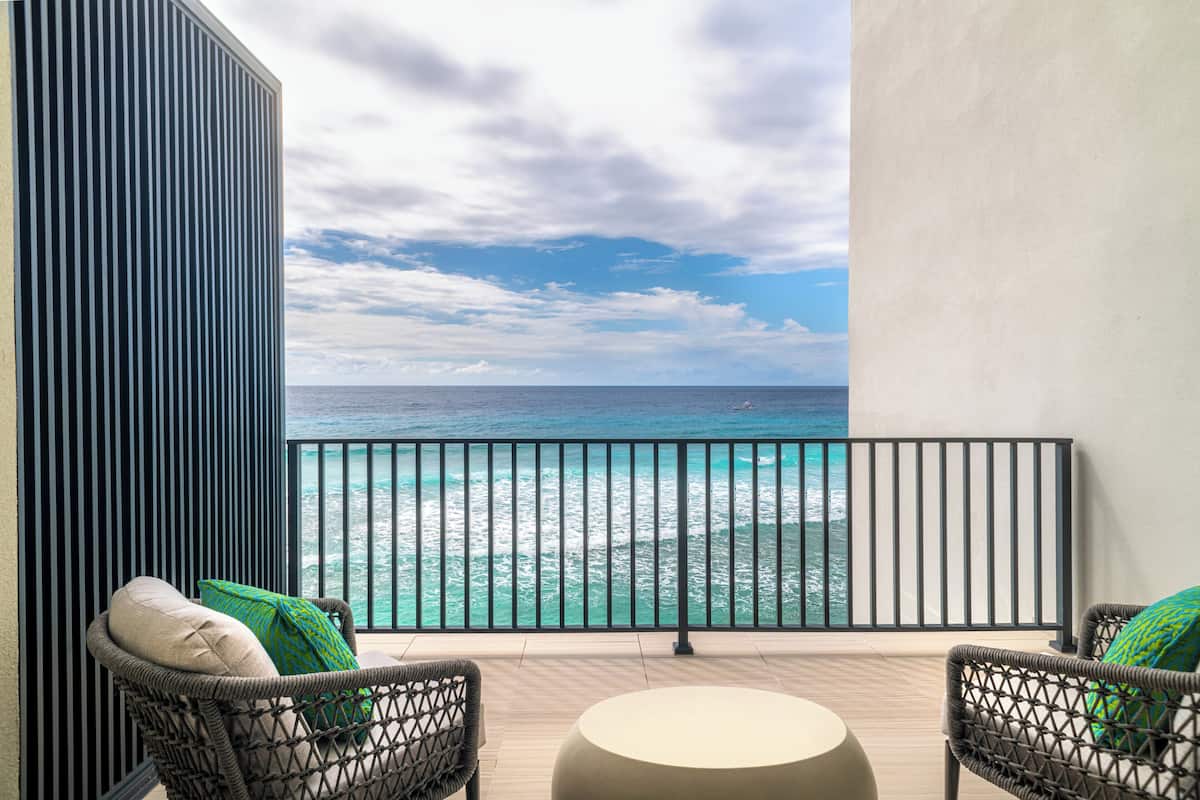 A balcony with wicker chairs and a round table, overlooking a turquoise ocean with waves.