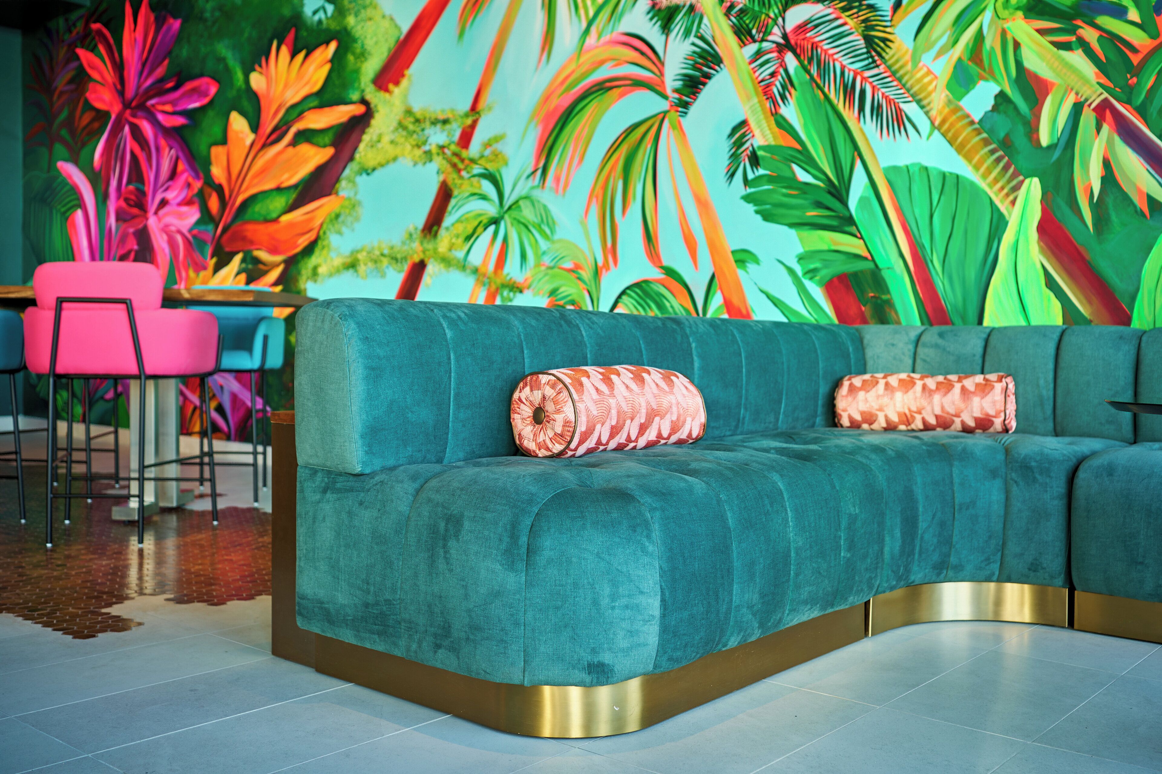 A teal sofa with gold trim, two patterned cushions, and a vibrant tropical mural in the background.