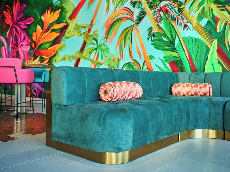A teal sofa with gold trim, two patterned cushions, and a vibrant tropical mural in the background.