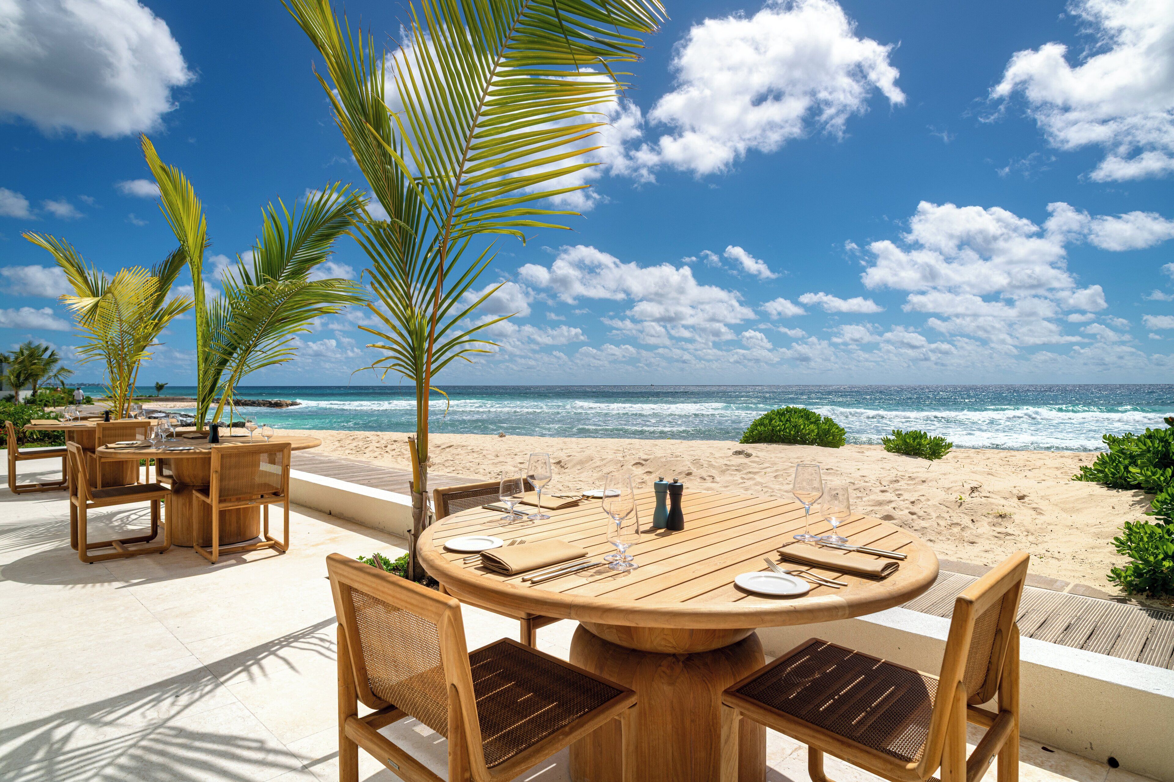 Ein Esszimmer mit Holztisch und Stühlen, mit Blick auf einen Strand mit Palmen und dem Meer.