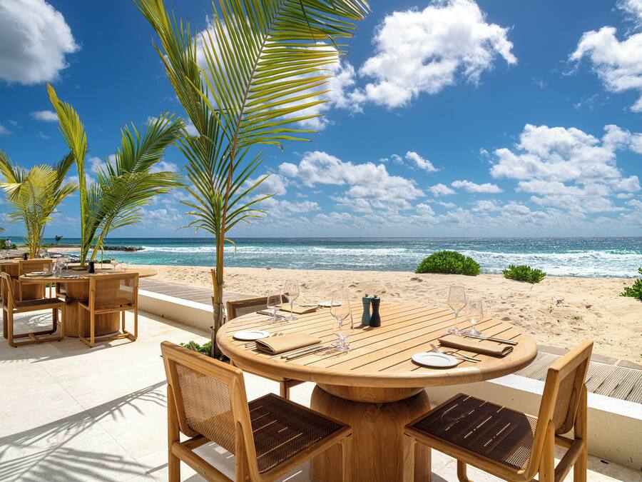Mesa de madera dispuesta para comer, con sillas, con vistas a una playa con palmeras y el océano.