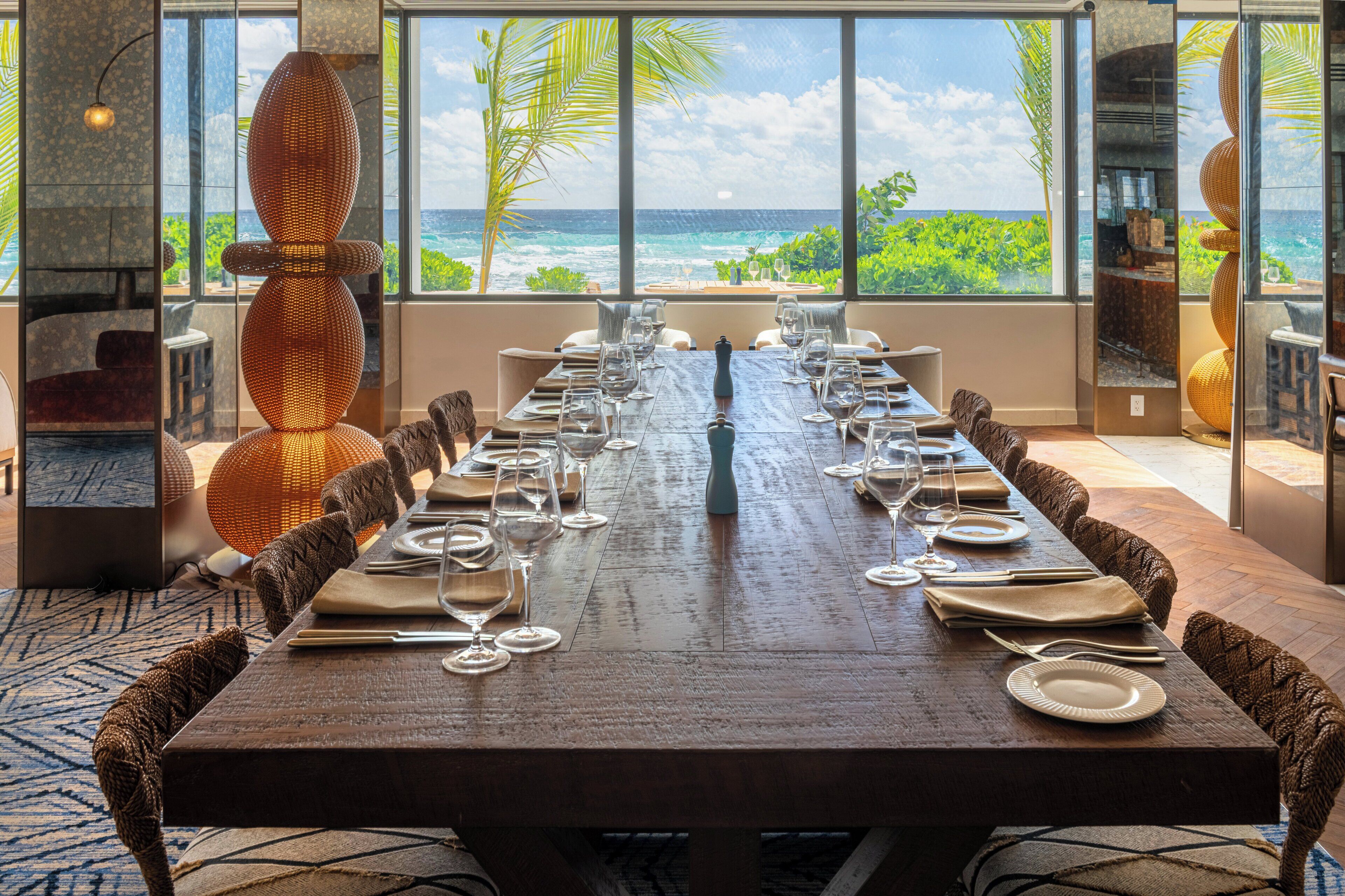 A dining area with a long table set for a meal, large windows with a view of the ocean, and tropical plants.