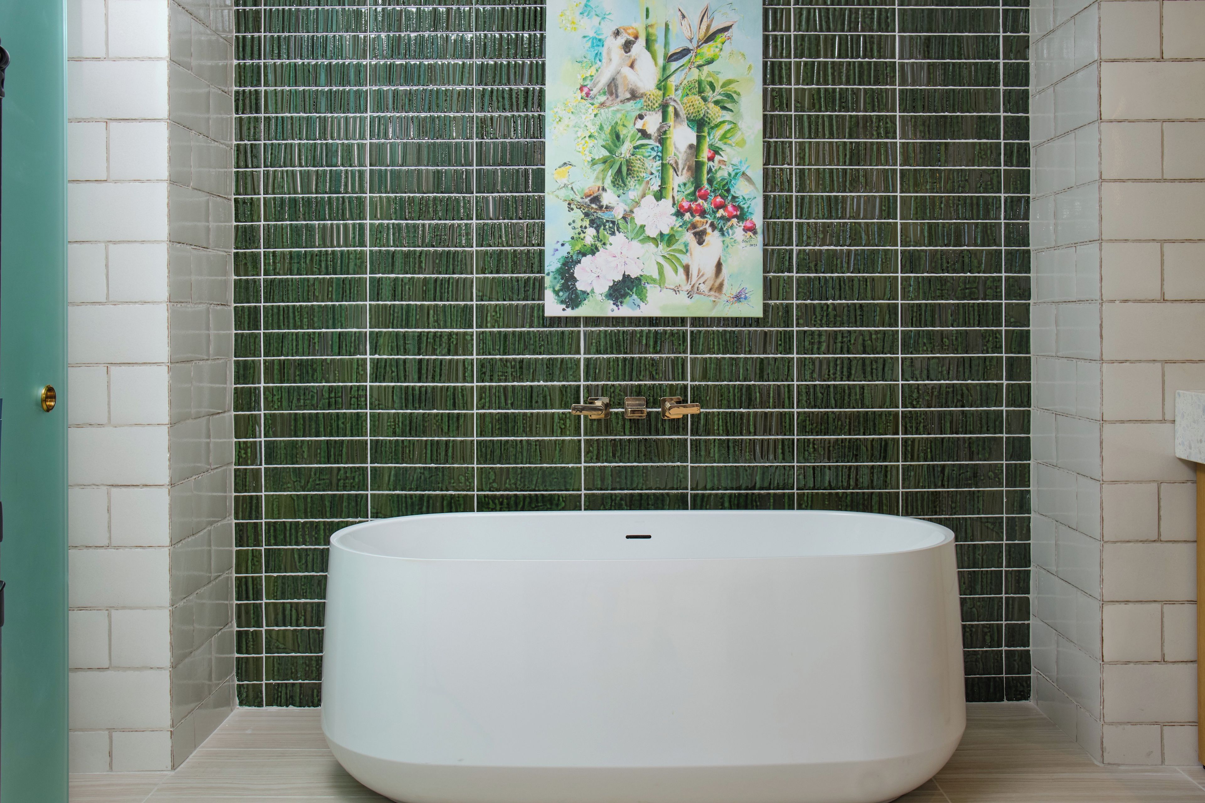 A bathroom with a large white bathtub, green mosaic tiles, and a painting of monkeys and flowers on the wall.