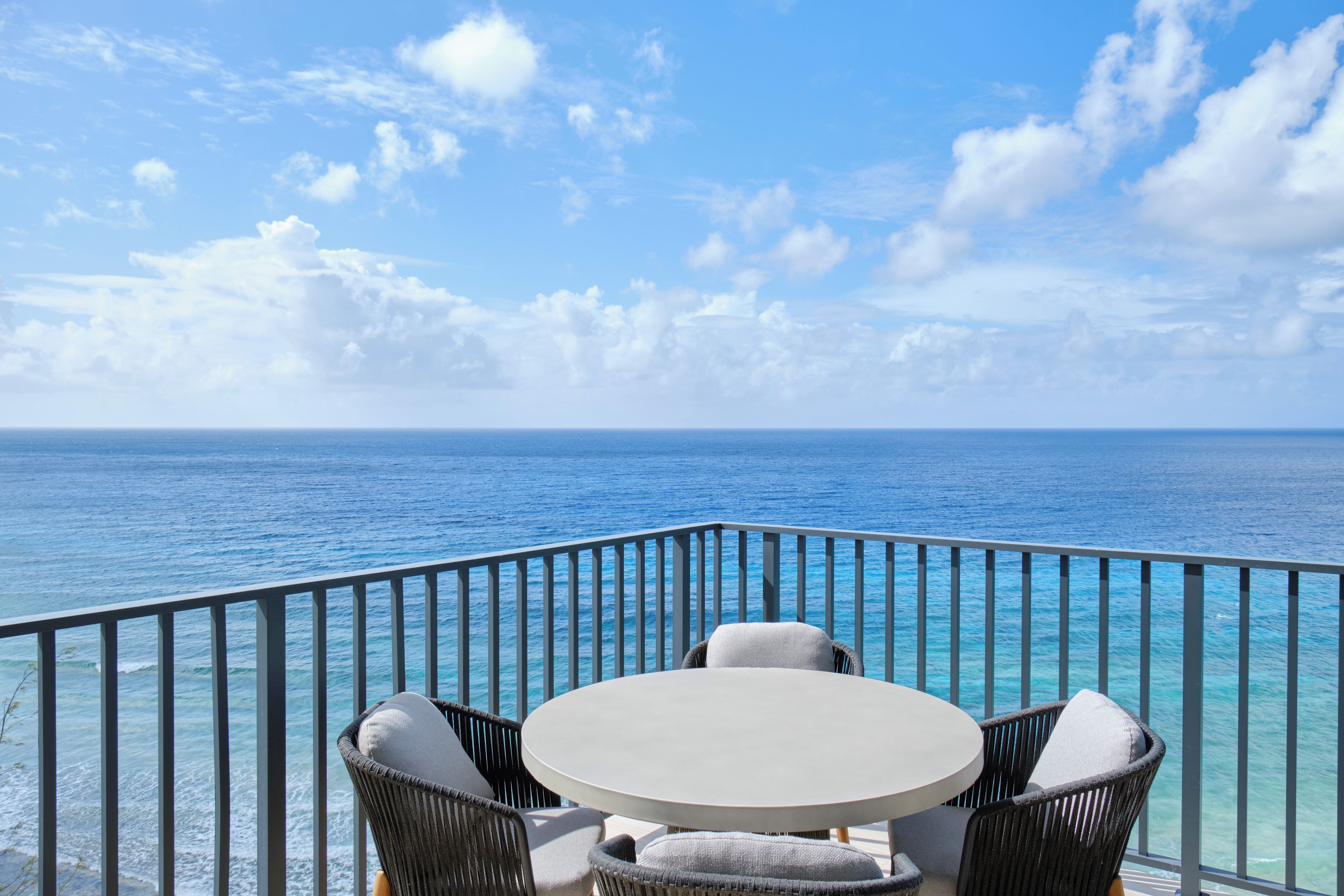 Varanda com mesa redonda e duas cadeiras, com vista para um mar azul e límpido.