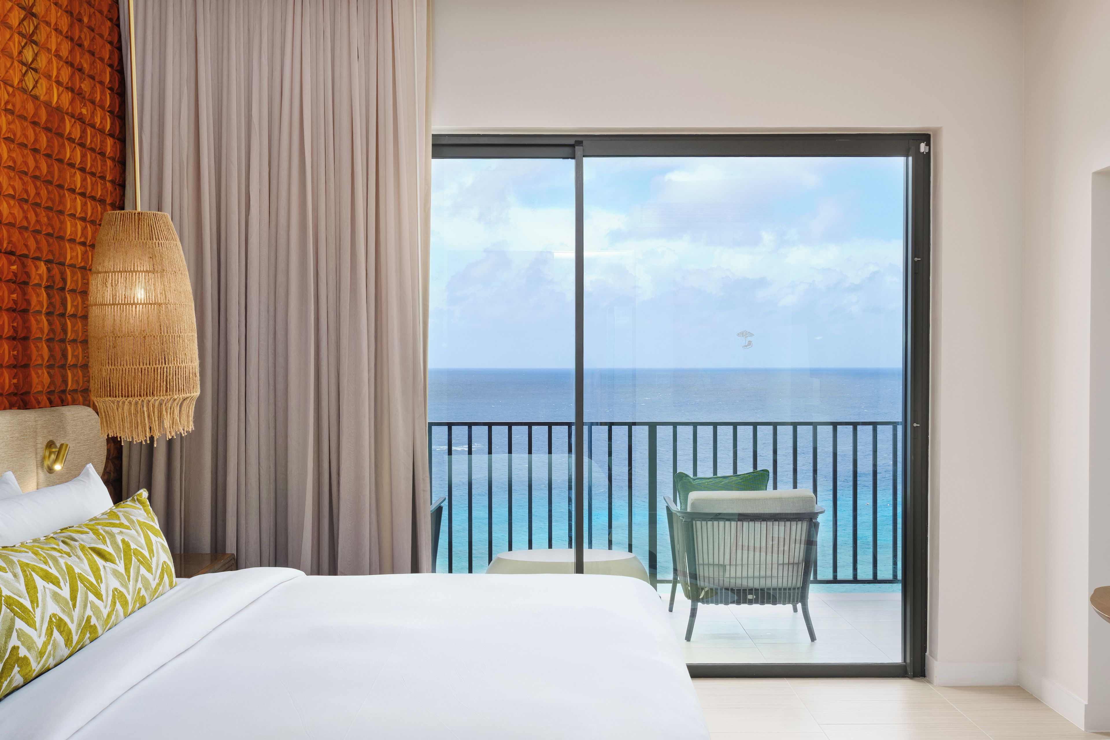 Habitación de hotel con cama, una silla, balcón con vista al mar y un ventanal grande.