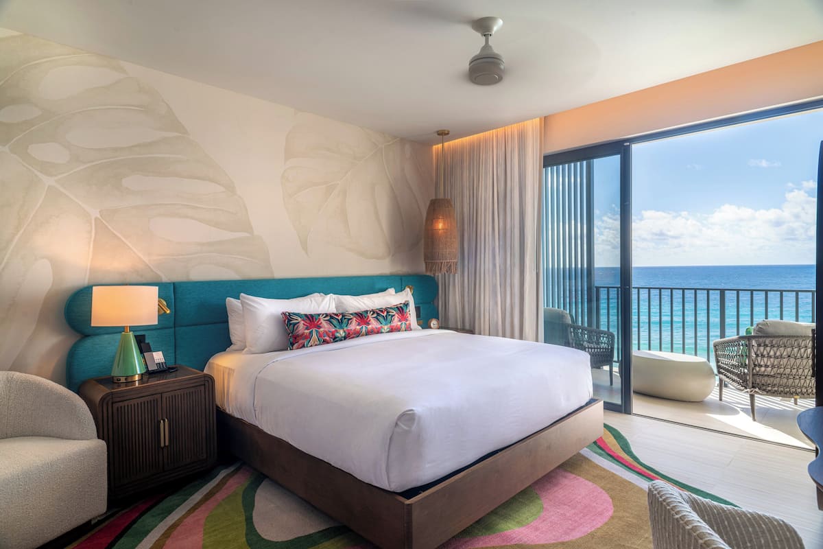 A hotel room with a large bed, a nightstand, a chair, a balcony with a view of the ocean.