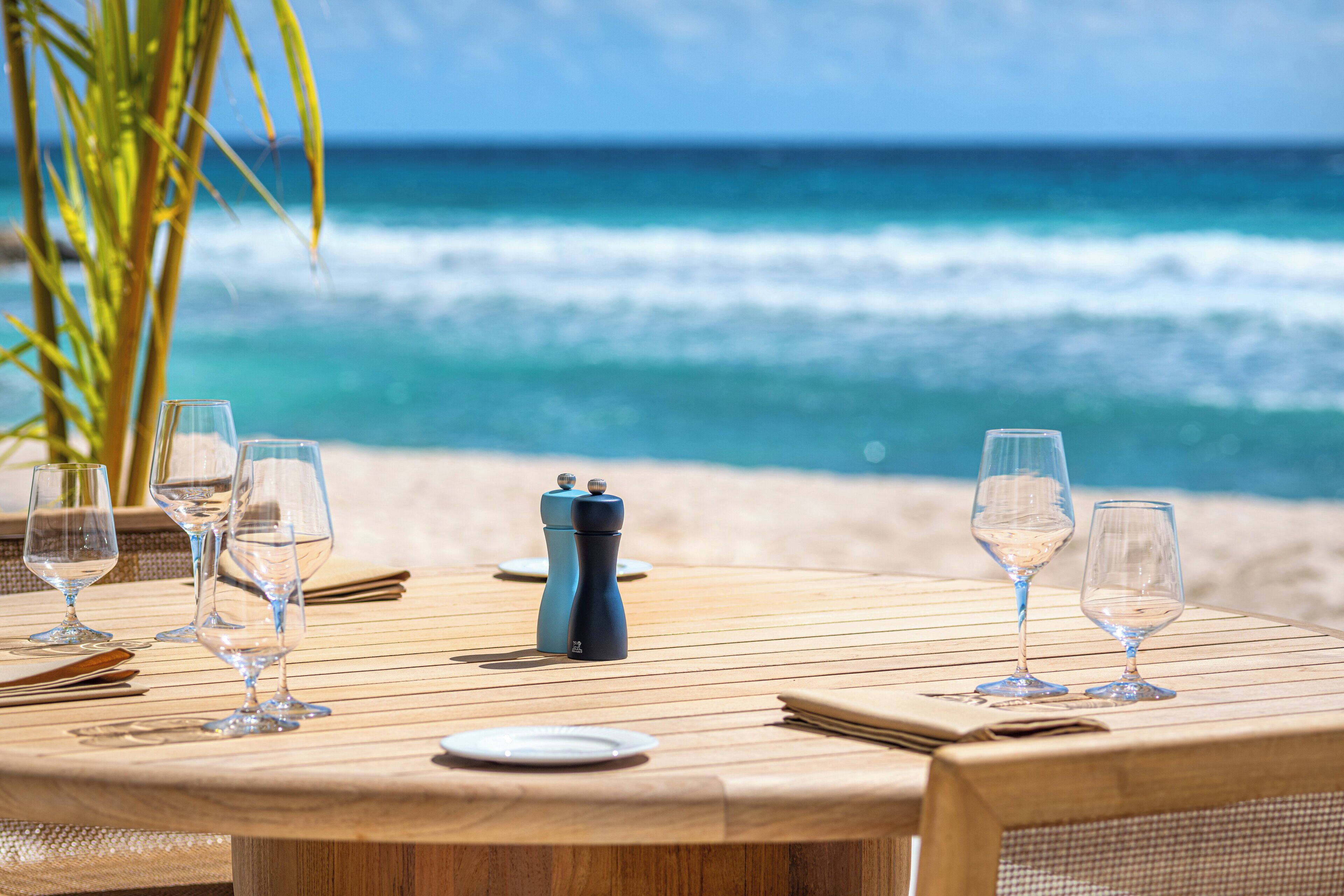 A wooden table set for dining with wine glasses and a salt and pepper shaker, overlooking a beach with waves.