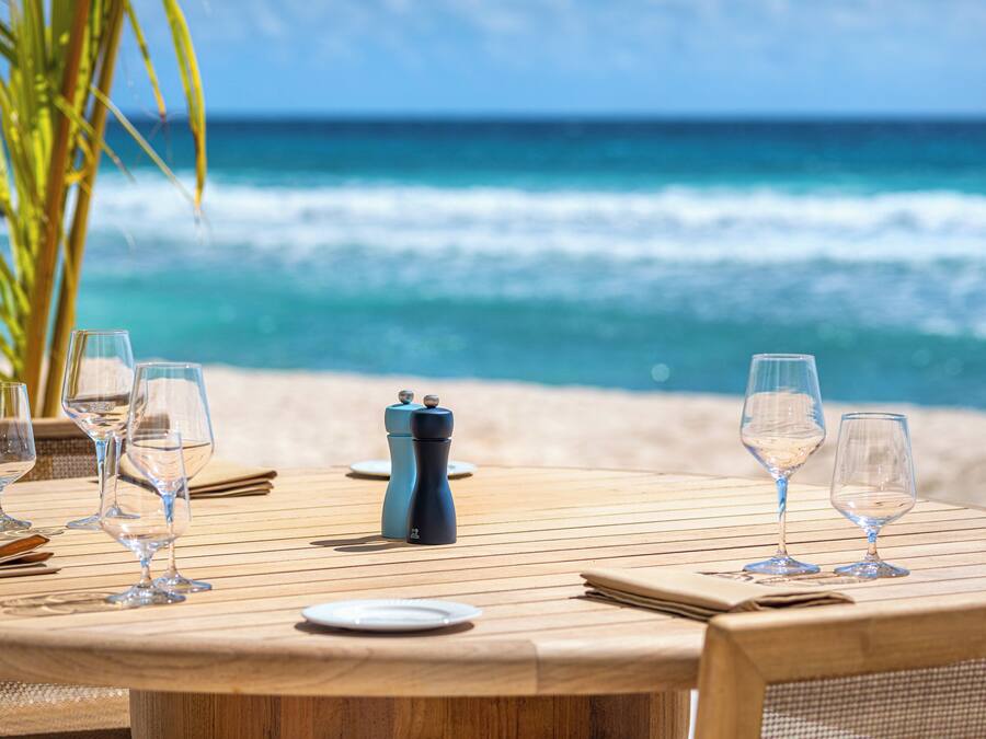 A wooden table set for dining with wine glasses and a salt and pepper shaker, overlooking a beach with waves.