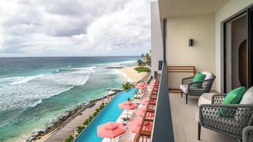 A balcony with a view of the ocean, a pool with umbrellas, and wicker furniture.