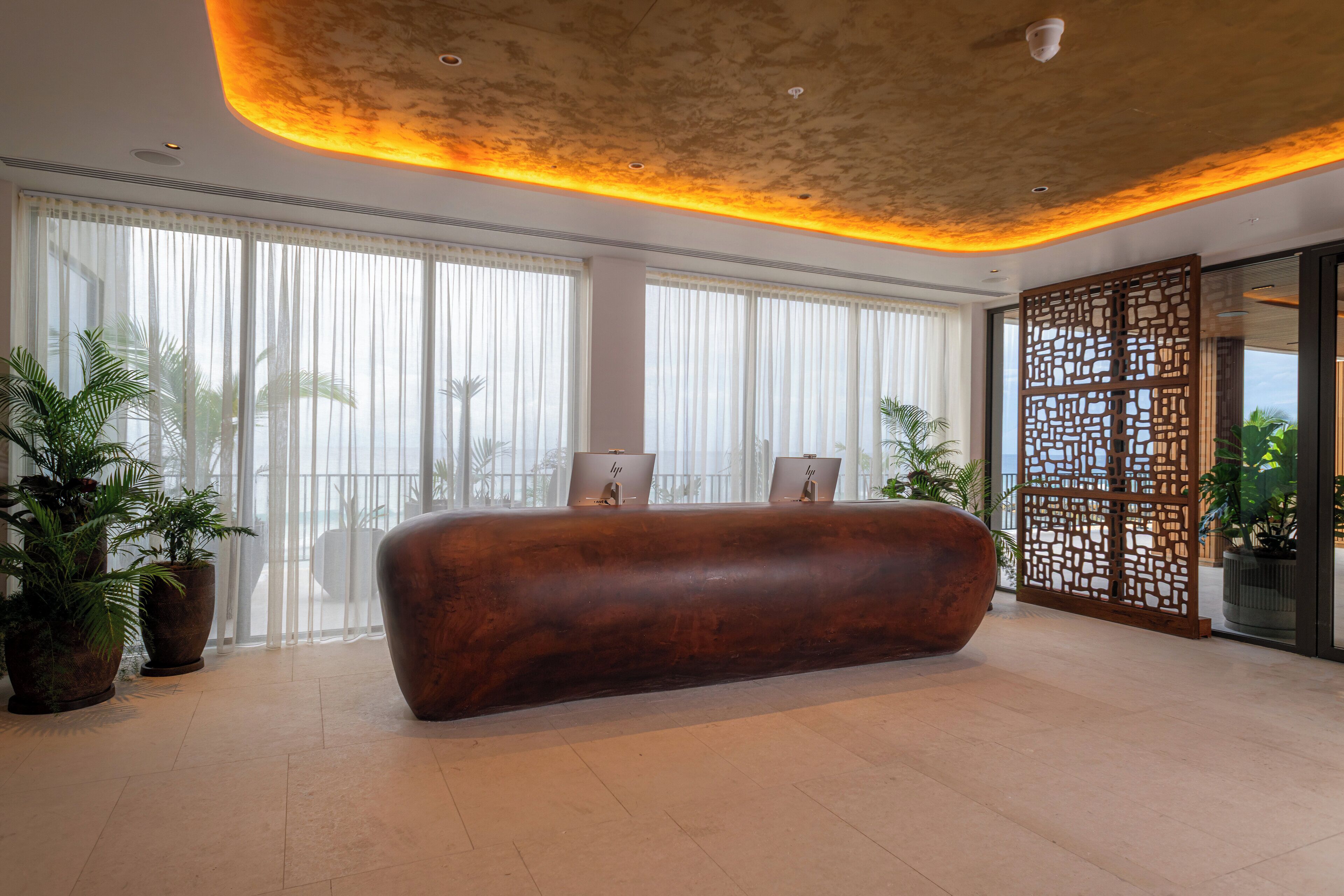 Un vestíbulo amplio con un mostrador de recepción grande, ventanas amplias con cortinas translúcidas y plantas en macetas.