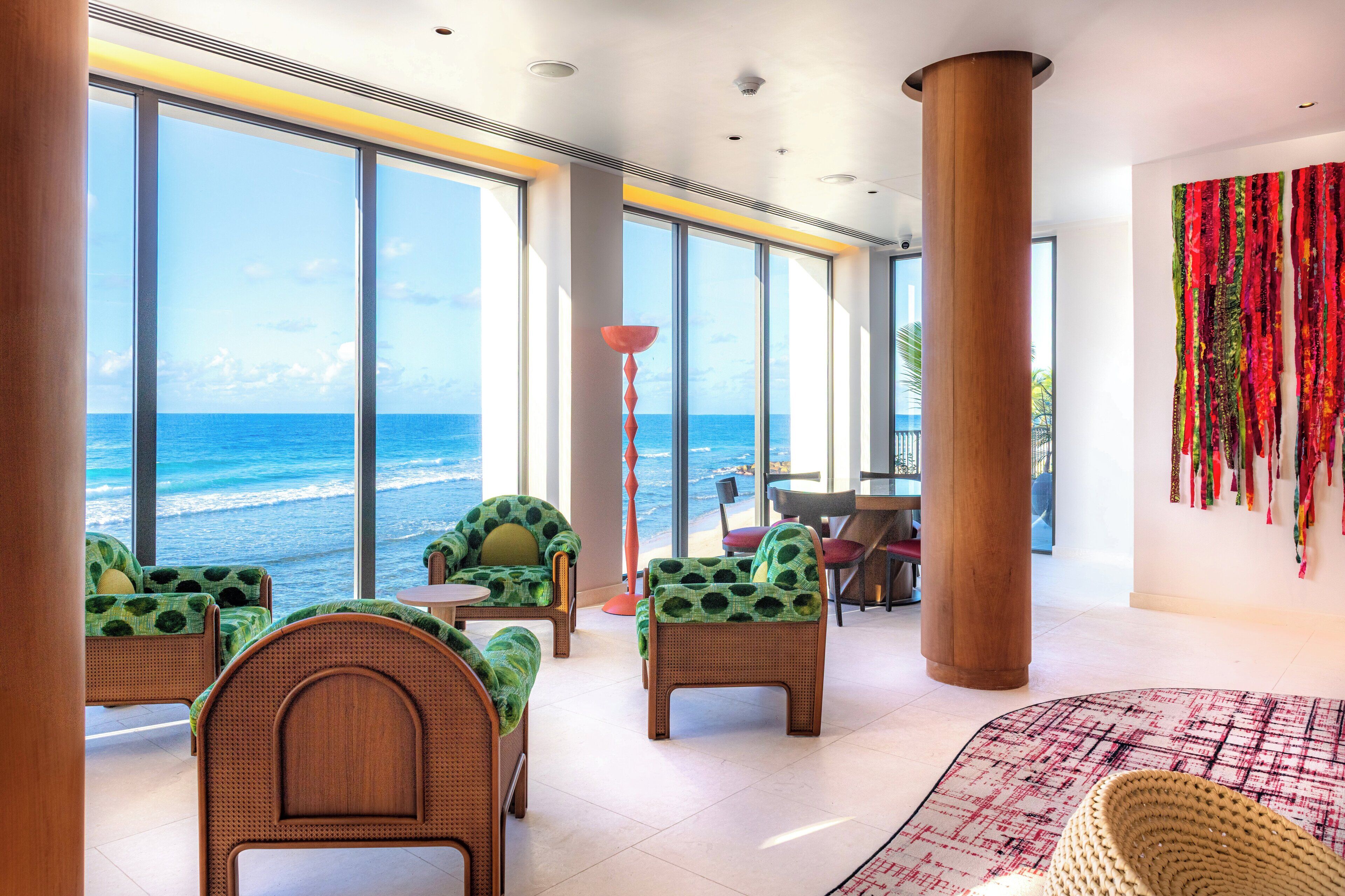 A spacious room with large windows overlooking the ocean, featuring wicker furniture and a red and white patterned rug.