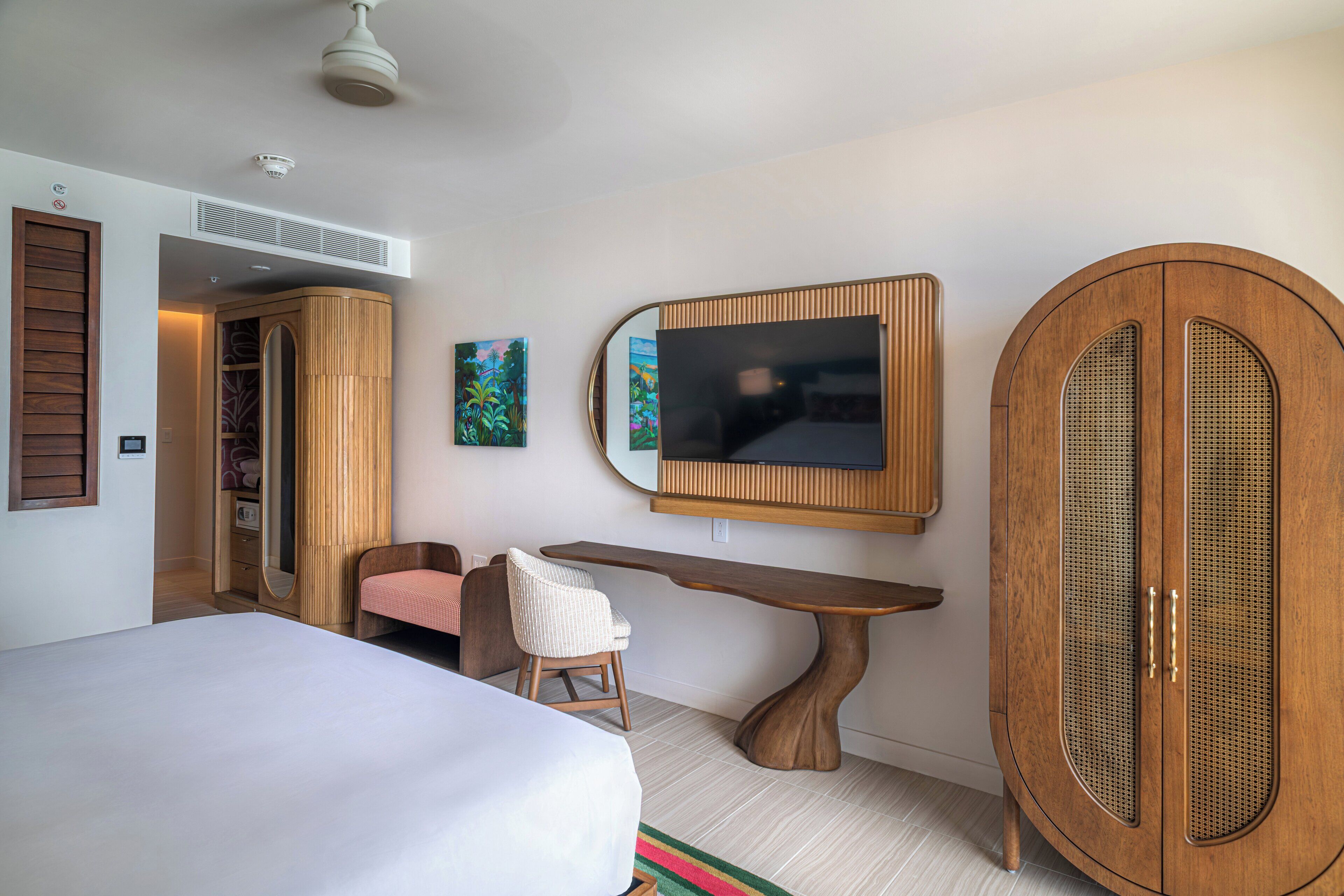 A hotel room with a bed, a wooden wardrobe, a desk, a chair, and a television mounted on the wall.