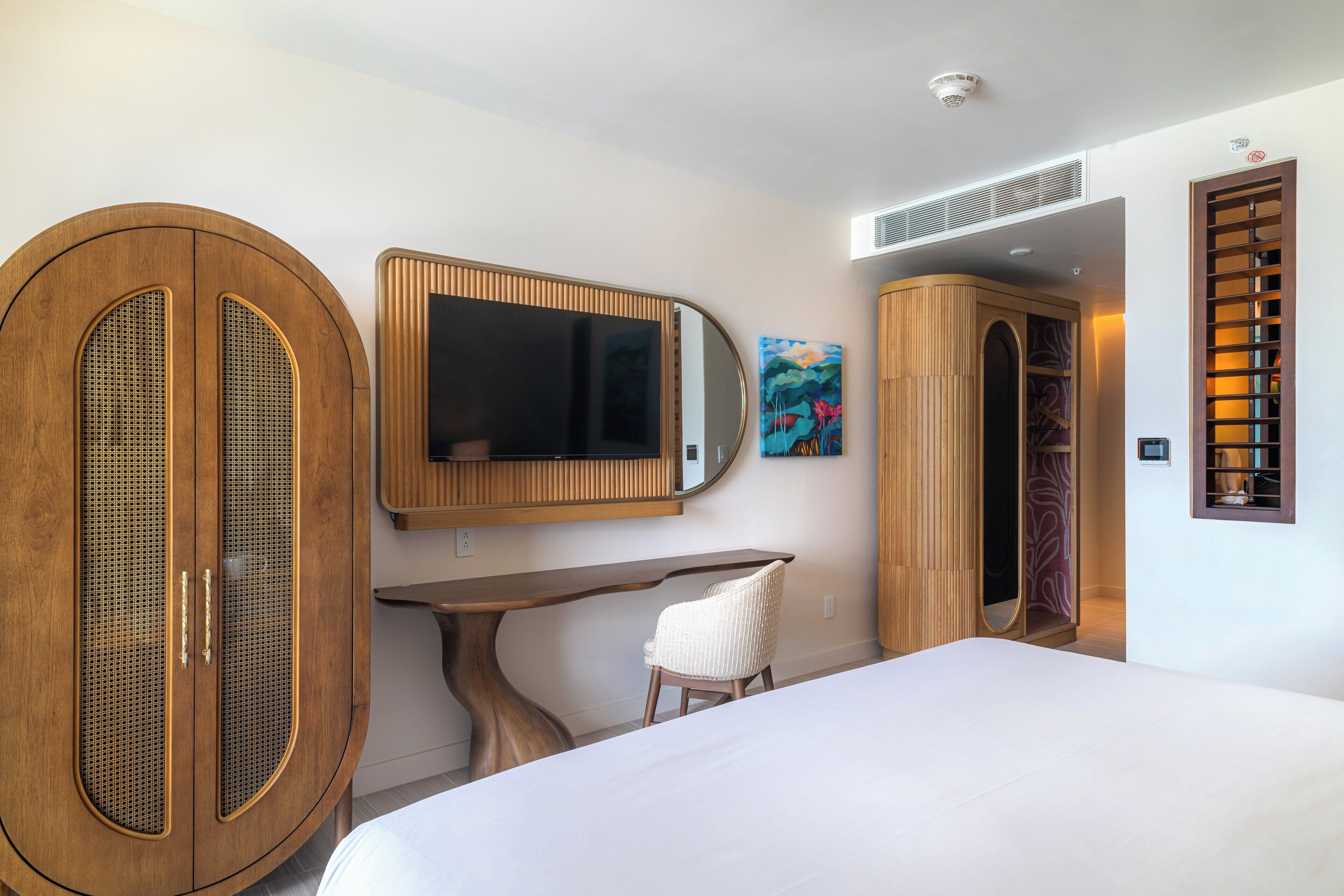 A bedroom with a bed, a wooden wardrobe, a desk, a chair, a large mirror, and a bookshelf.