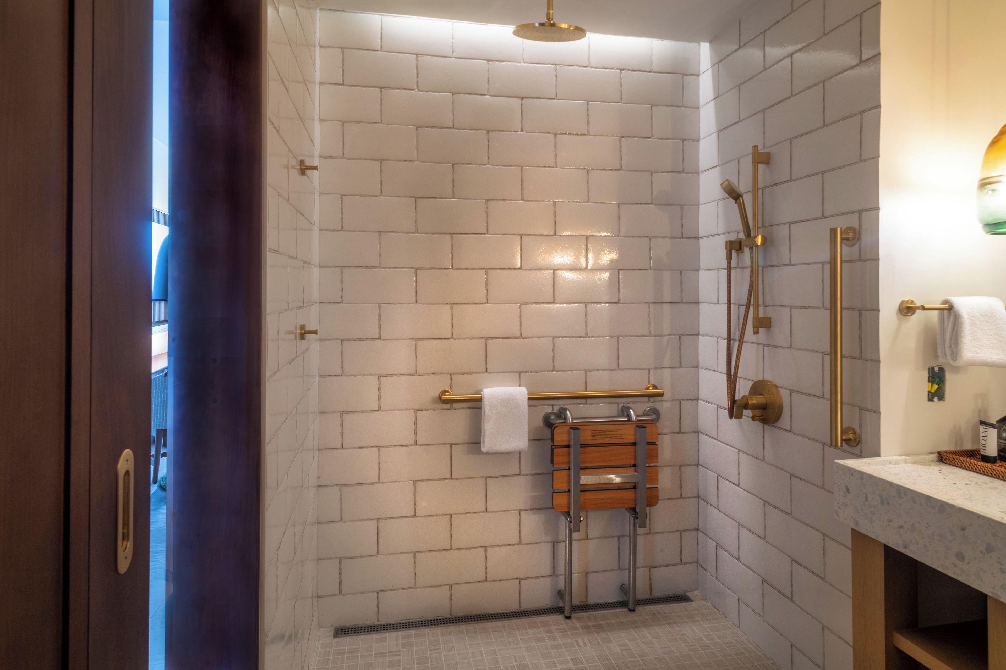 A modern bathroom with white brick walls, a wooden towel rack, a shower with a gold showerhead, and a vanity with a marble countertop.
