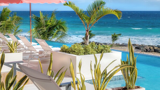 A poolside area with lounge chairs and a pink umbrella, overlooking a clear blue ocean. - Hotel Indigo Bridgetown Barbados by IHG (Hastings)
