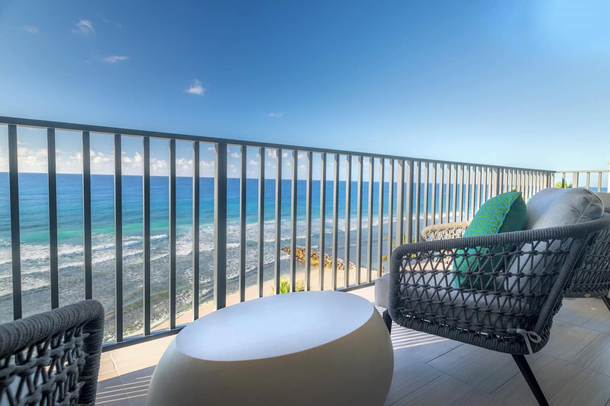 A balcony with wicker chairs and a round table, overlooking a beach with the ocean and a clear sky.
