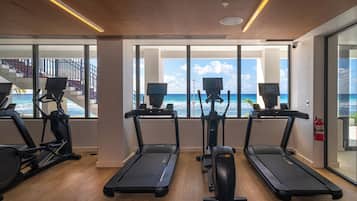 A modern gym with treadmills, elliptical machines, and stationary bikes. It features large windows with a view of the ocean and a staircase leading to an outdoor area.