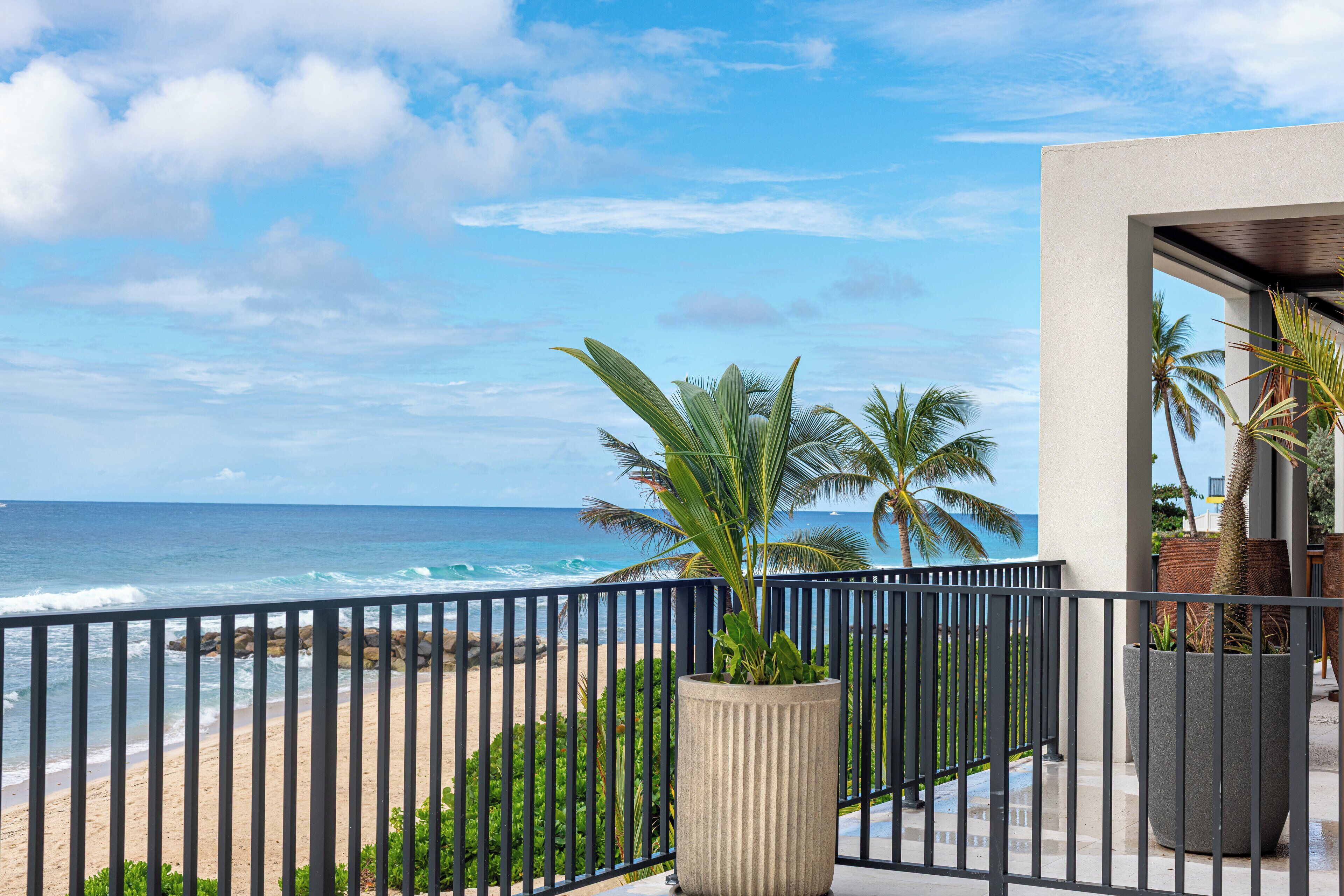 A balcony with a view of the ocean, palm trees, and a modern building with a concrete facade.