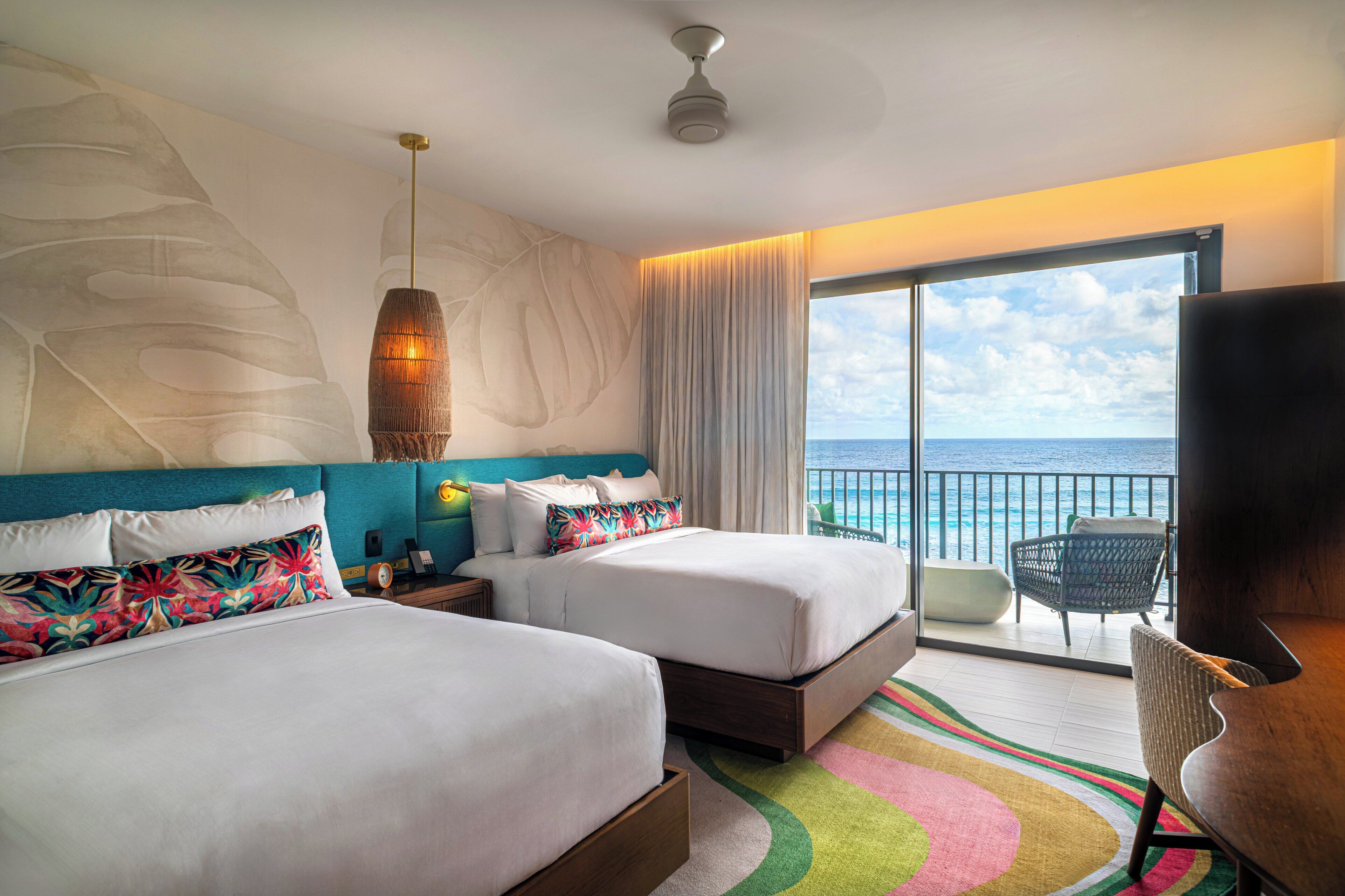 Une chambre d’hôtel avec deux lits, une grande fenêtre donnant sur l’océan, un ventilateur de plafond et un balcon avec une chaise.