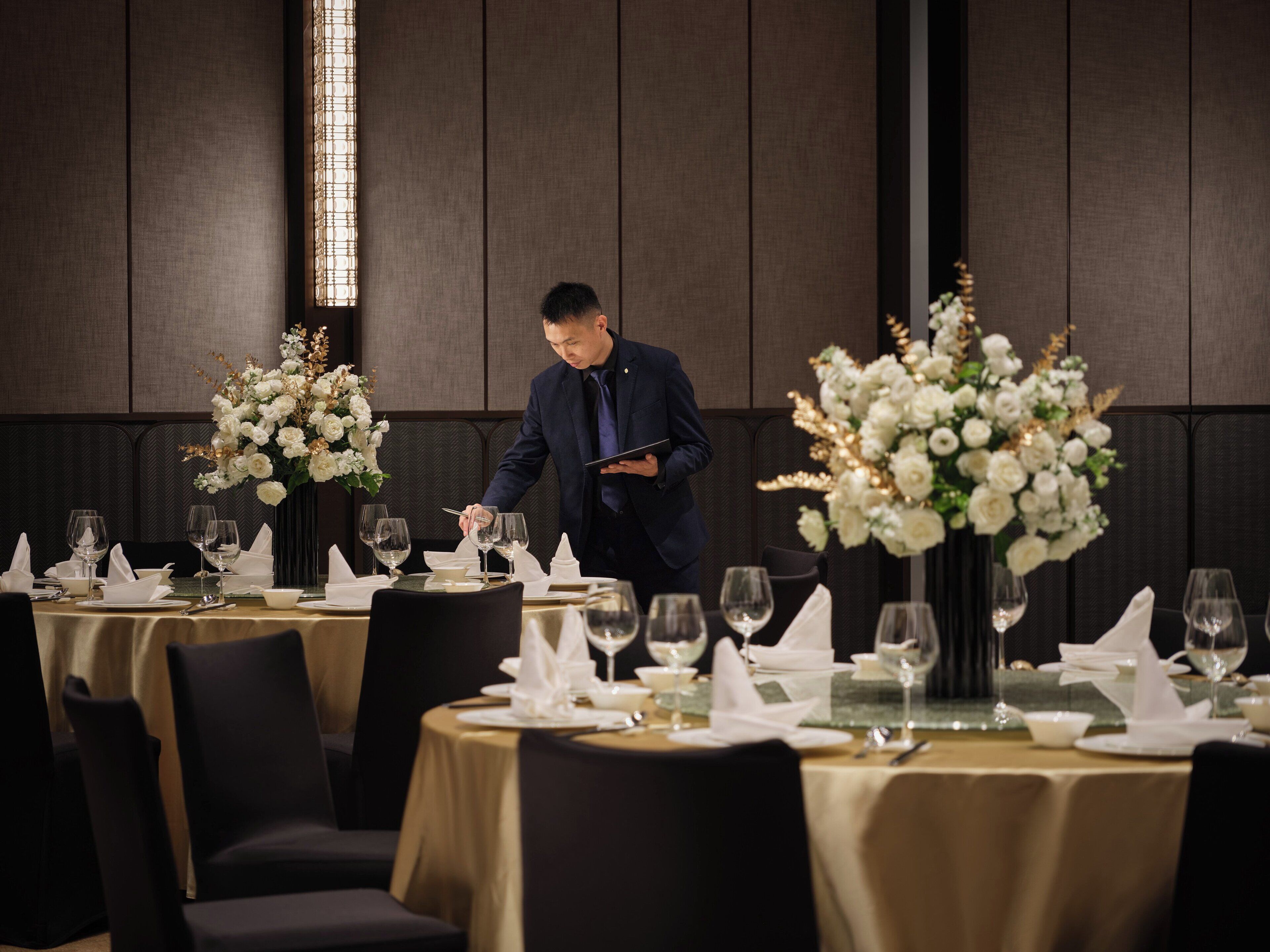 Un ambiente de restaurante con mesas dispuestas para comer, un camarero atendiendo a una mesa y grandes centros de flores.