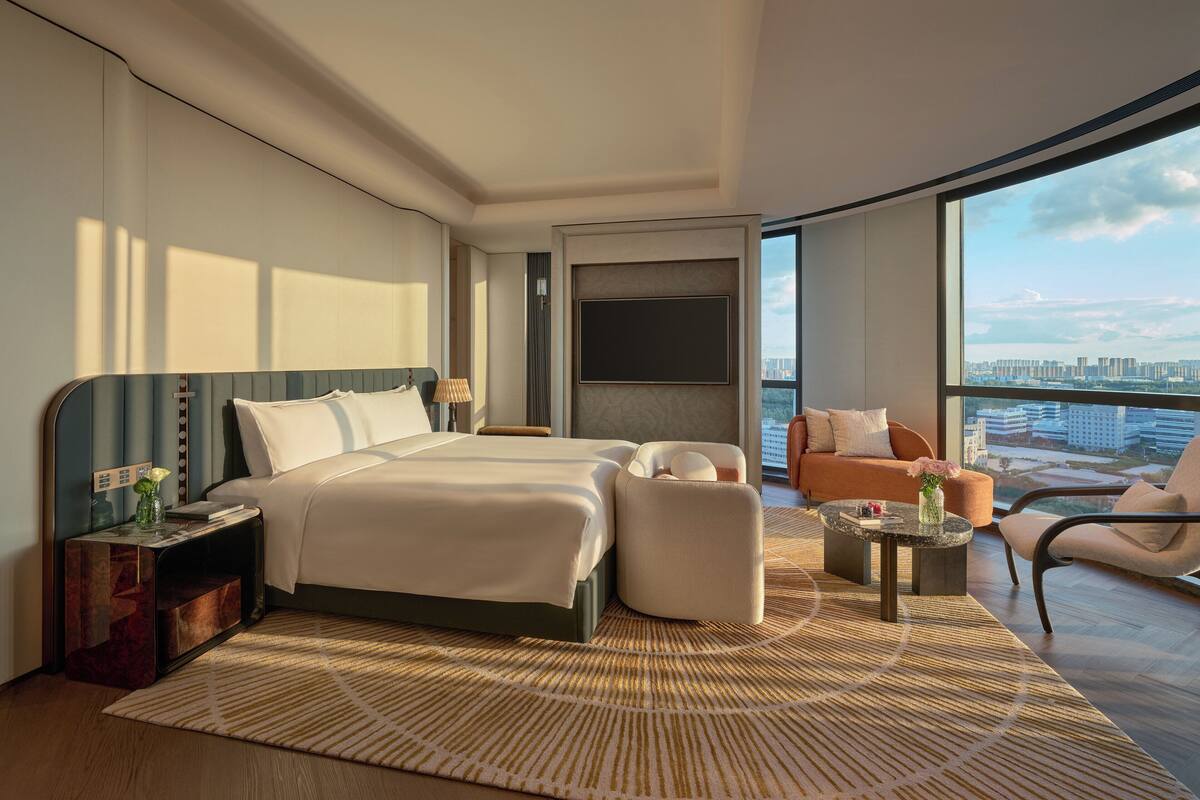 A modern hotel room with a large bed, two seating areas, a TV, and a city view.