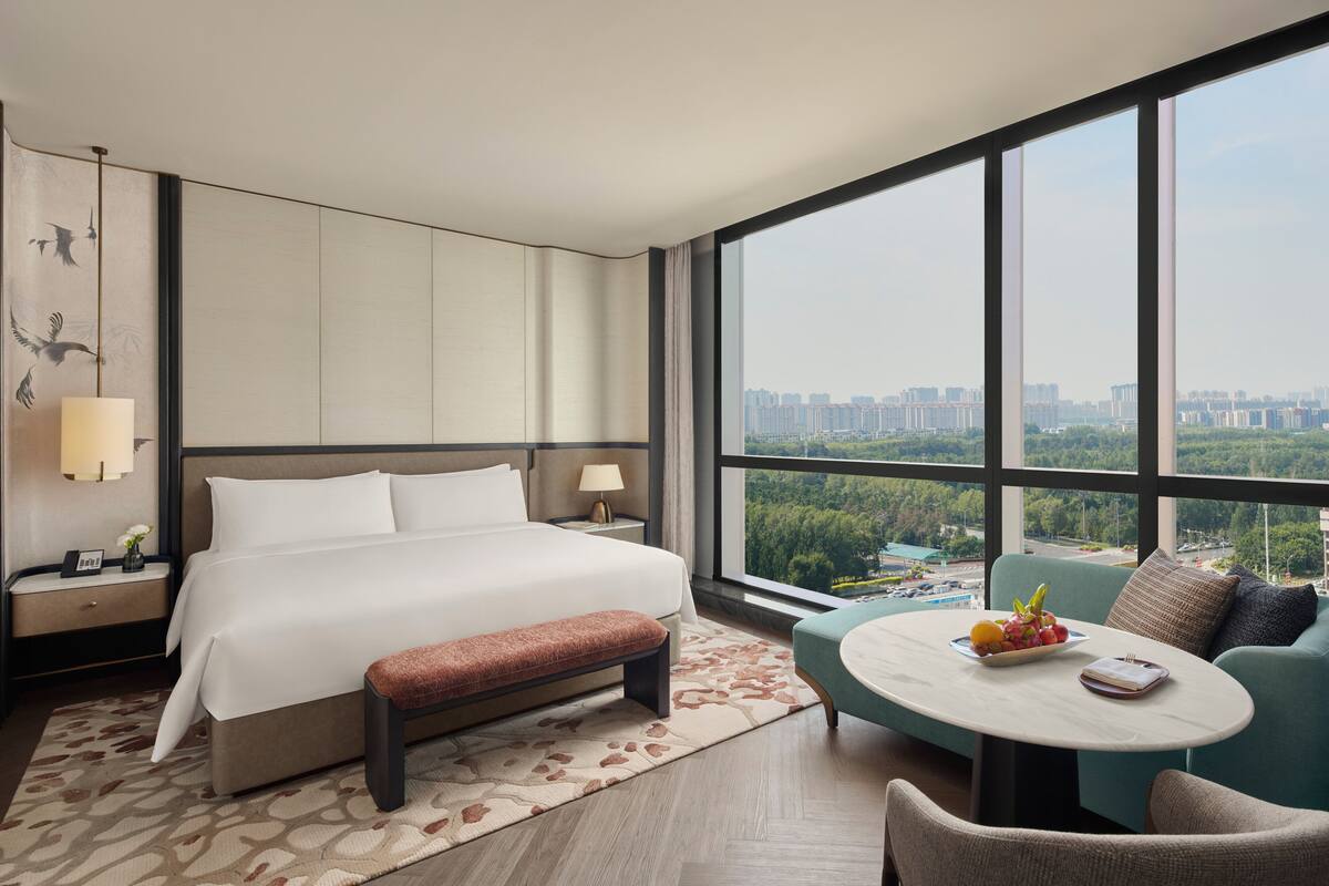 A modern hotel room with a large bed, a bench, a round table with fruit, and a view of a cityscape through large windows.