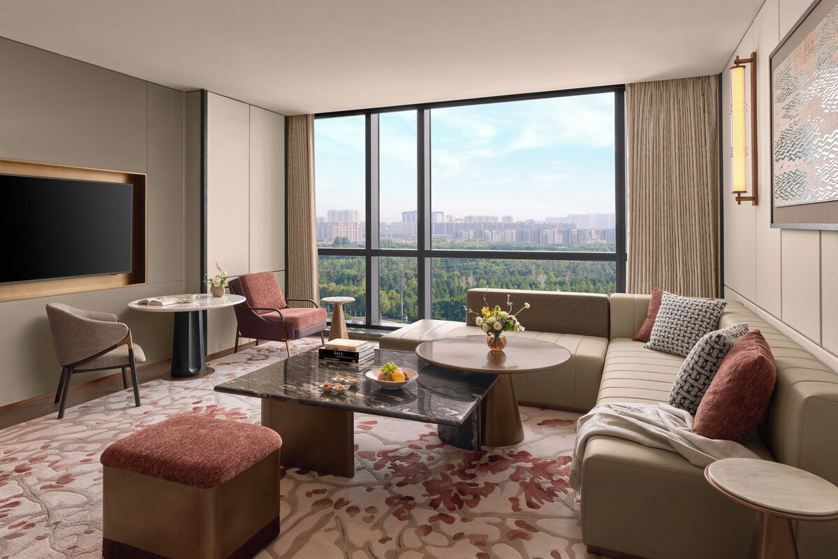 Habitación de hotel moderna con un ventanal grande, un televisor de pantalla plana, un sofá, una mesa de centro y una silla.